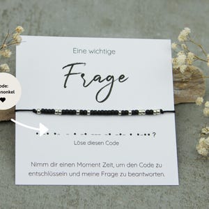 Könnte beinhalten: Eine weiße Karte mit schwarzem Text, der "Code: Patenonkel" und "Eine wichtige Frage" lautet, mit einem schwarzen und silbernen Perlenarmband. Die Karte enthält auch den Text "Löse diesen Code" und eine Reihe von Punkten und Strichen, gefolgt von einem Fragezeichen. Die Karte endet mit dem Text "Nimm dir einen Moment Zeit, um den Code zu entschlüsseln und meine Frage zu beantworten."