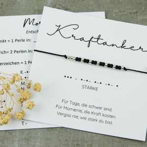 May include: A black beaded bracelet with silver accents is displayed on a white card with the word "Kraftanker" in script. The card also has text in German, including the word "St&auml;rke" and a Morse code pattern. Dried flowers are in the foreground.
