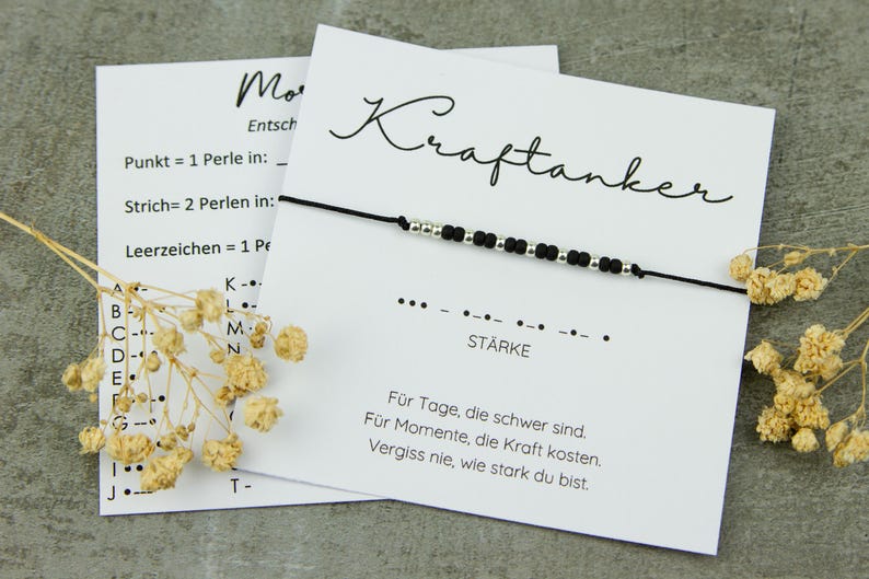 May include: A black beaded bracelet with silver accents is displayed on a white card with the word "Kraftanker" in script. The card also has text in German, including the word "St&auml;rke" and a Morse code pattern. Dried flowers are in the foreground.