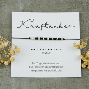 May include: A black cord bracelet with silver beads and black beads, displayed on a white card with the German word "Kraftanker" and the word "ST&Auml;RKE". The card also includes text in German and is decorated with dried flowers.