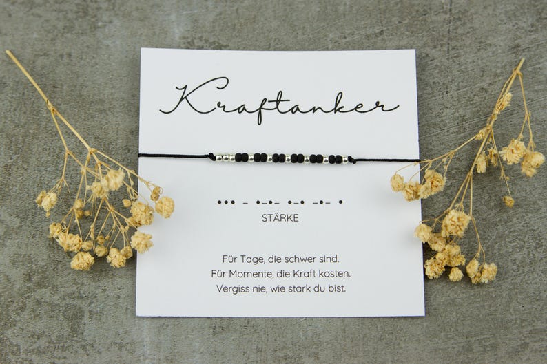 May include: A black cord bracelet with silver beads and black beads, displayed on a white card with the German word "Kraftanker" and the word "ST&Auml;RKE". The card also includes text in German and is decorated with dried flowers.