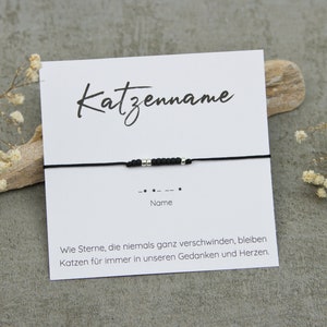 Könnte beinhalten: Ein schwarzes Armband mit silbernen Perlen und einer weißen Karte. Auf der Karte steht das Wort "Katzenname" in Schreibschrift, das Wort "Name" und ein Morsecode-Muster. Die Karte enthält auch Text auf Deutsch.