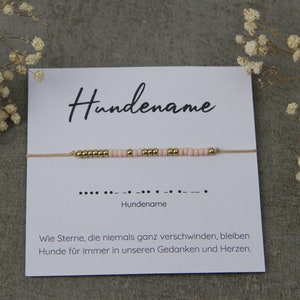 Könnte beinhalten: Eine weiße Karte mit dem Text "Hundename" und einem beigefarbenen Armband aus Schnur mit rosa und goldenen Perlen. Die Karte enthält auch den Text "Wie Sterne, die niemals ganz verschwinden, bleiben Hunde für immer in unseren Gedanken und Herzen."