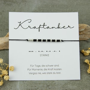 Könnte beinhalten: Ein schwarzes Kordelarmband mit silbernen und schwarzen Perlen, präsentiert auf einer weißen Karte. Die Karte zeigt das Wort "Kraftanker" in Schrift, Morsecode und das Wort "STÄRKE".