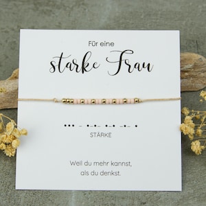 Könnte beinhalten: Ein zartes Armband mit rosa und goldenen Perlen auf einer dünnen Schnur, präsentiert auf einer weißen Karte mit deutschem Text. Die Karte trägt die Aufschrift "Für eine starke Frau" und "Stärke" mit einem Morsecode-Muster. Trockenblumen und Holzakzente umgeben die Karte.