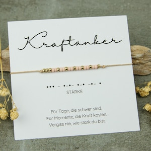 Könnte beinhalten: Ein zartes Armband mit rosa und goldenen Perlen auf einer dünnen Schnur, präsentiert auf einer Karte mit dem Wort "Kraftanker". Die Karte enthält auch deutschen Text und ein Morsecode-Muster.