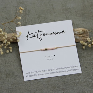 Könnte beinhalten: Eine weiße Karte mit dem Text "Katzenname" und "Name" darauf gedruckt. Ein beigefarbenes Band mit rosa und goldenen Perlen ist an der Karte befestigt. Der Text unter den Perlen lautet "Wie Sterne, die niemals ganz verschwinden, bleiben Katzen für immer in unseren Gedanken und Herzen."