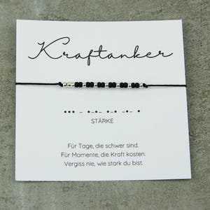 May include: A black string bracelet with silver and black beads is displayed on a white card. The card has the word "Kraftanker" in cursive, Morse code, and the word "ST&Auml;RKE". Text below reads: "For days that are difficult. For moments that cost strength. Never forget how strong you are."