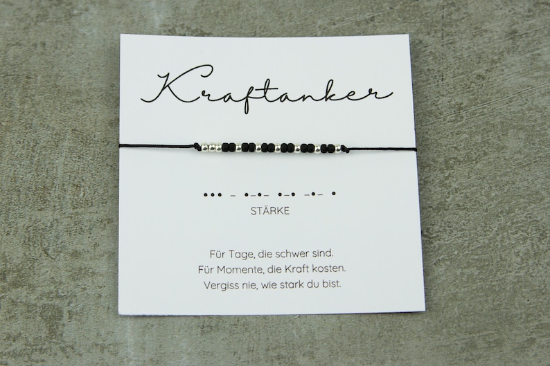 May include: A black string bracelet with silver and black beads is displayed on a white card. The card has the word "Kraftanker" in cursive, Morse code, and the word "ST&Auml;RKE". Text below reads: "For days that are difficult. For moments that cost strength. Never forget how strong you are."