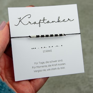 May include: A black string bracelet with silver and black beads is displayed on a white card. The card has the word "Kraftanker" in cursive, Morse code, and the word "ST&Auml;RKE". The card also has text in German.
