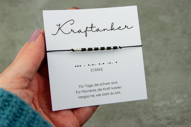 May include: A black string bracelet with silver and black beads is displayed on a white card. The card has the word "Kraftanker" in cursive, Morse code, and the word "ST&Auml;RKE". The card also has text in German.