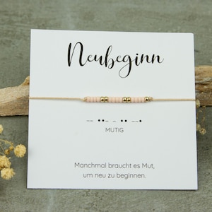 Könnte beinhalten: Ein zartes Armband mit rosa und goldenen Perlen auf einer beigen Schnur, präsentiert auf einer weißen Karte. Die Karte zeigt das Wort "Neubeginn" in eleganter Schrift sowie das Wort "MUTIG" in Morsecode. Die Karte hat auch eine Nachricht auf Deutsch.
