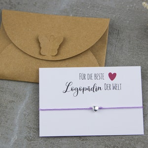 Könnte beinhalten: Eine weiße Karte mit dem Text "Für die beste Logopädin der Welt" und einem roten Herz. Ein lilafarbenes Armband aus Schnur mit einem silbernen Herz-Anhänger liegt auf der Karte. Die Karte liegt auf einem braunen Umschlag mit einem Schmetterlingsdesign.