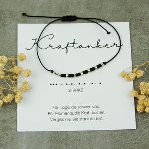 May include: A black cord bracelet with silver beads and a sliding knot closure. The bracelet is displayed on a white card with the word "Kraftanker" and the word "ST&Auml;RKE" printed on it. The card also has a message in German.