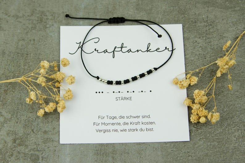 May include: A black cord bracelet with silver beads and a sliding knot closure. The bracelet is displayed on a white card with the word "Kraftanker" and the word "ST&Auml;RKE" printed on it. The card also has a message in German.