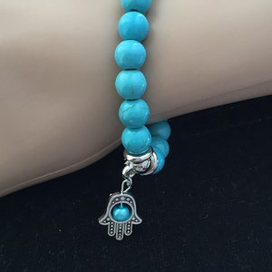 May include: A turquoise beaded bracelet with a silver hamsa charm. The hamsa charm has a turquoise stone in the center.