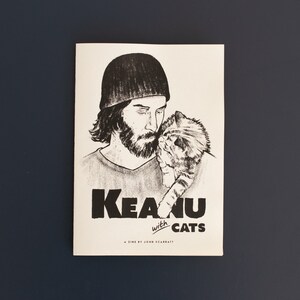Pode incluir: Ilustra&ccedil;&atilde;o a preto e branco de uma pessoa com barba e um gato sobre fundo branco. O texto "Keanu with cats" est&aacute; impresso em negrito preto. A revista est&aacute; sobre uma superf&iacute;cie azul escura.