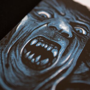 Puede incluir: Una impresi&oacute;n gr&aacute;fica en negro y azul que muestra una ilustraci&oacute;n en primer plano de una criatura gru&ntilde;endo con los dientes descubiertos y los ojos muy abiertos. La obra utiliza sombras de alto contraste para definir los rasgos faciales.