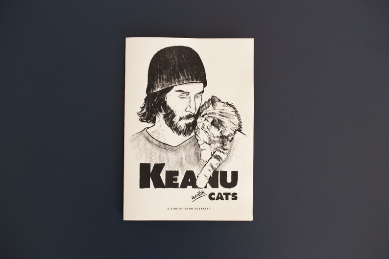 Pode incluir: Ilustra&ccedil;&atilde;o a preto e branco de uma pessoa com barba e um gato sobre fundo branco. O texto "Keanu with cats" est&aacute; impresso em negrito preto. A revista est&aacute; sobre uma superf&iacute;cie azul escura.