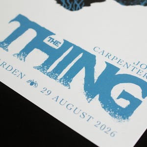 Puede incluir: Un p&oacute;ster blanco con el texto azul THE THING, John Carpenter's, y el texto STATION GARDEN 29 AUGUST 2026 junto a un peque&ntilde;o icono de ara&ntilde;a. El dise&ntilde;o incluye un elemento gr&aacute;fico oscuro y texturizado en el borde superior del papel blanco.