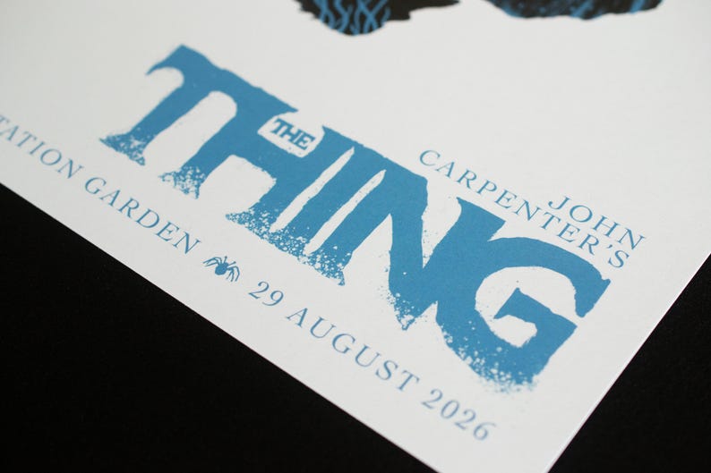 Puede incluir: Un p&oacute;ster blanco con el texto azul THE THING, John Carpenter's, y el texto STATION GARDEN 29 AUGUST 2026 junto a un peque&ntilde;o icono de ara&ntilde;a. El dise&ntilde;o incluye un elemento gr&aacute;fico oscuro y texturizado en el borde superior del papel blanco.