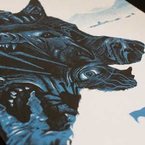 Puede incluir: Un p&oacute;ster serigrafiado con una ilustraci&oacute;n en azul oscuro y negro de una criatura de varias cabezas con dientes afilados y un ojo prominente. La obra incluye la silueta de un veh&iacute;culo a lo lejos y el texto azul parcial THE.