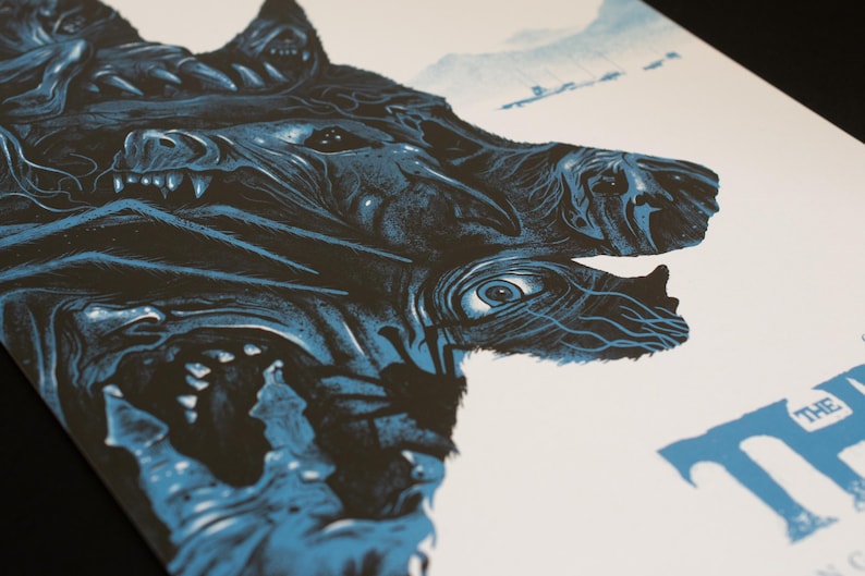 Puede incluir: Un p&oacute;ster serigrafiado con una ilustraci&oacute;n en azul oscuro y negro de una criatura de varias cabezas con dientes afilados y un ojo prominente. La obra incluye la silueta de un veh&iacute;culo a lo lejos y el texto azul parcial THE.