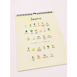 May include: A spiral-bound calendar with the word "Janvier" at the top. The calendar features colorful illustrations of various fruits and vegetables, including avocado, carrot, and kiwi. The calendar is on a light yellow background.