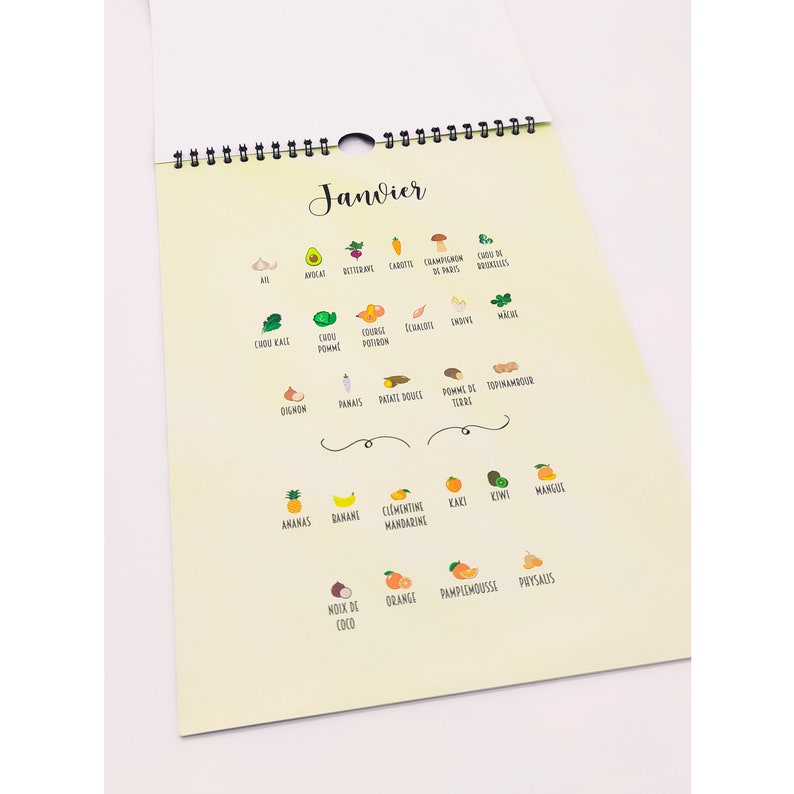 May include: A spiral-bound calendar with the word "Janvier" at the top. The calendar features colorful illustrations of various fruits and vegetables, including avocado, carrot, and kiwi. The calendar is on a light yellow background.