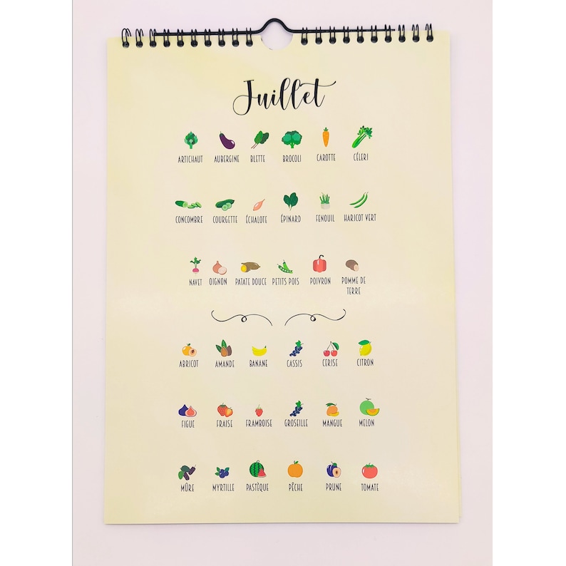 May include: A spiral-bound calendar page with the word "Juillet" at the top. It features colorful illustrations of various fruits and vegetables, including artichoke, eggplant, broccoli, carrot, and celery. The page is cream-colored.