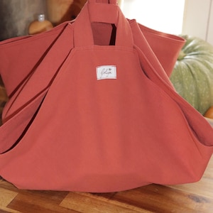 May include: A large, terracotta-colored tote bag with a rectangular label featuring cursive script. The bag has two handles and a wide opening. The bag is made of a soft, durable fabric, perfect for carrying groceries or other items.