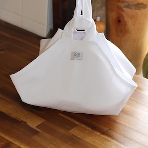 May include: A white tote bag with a rectangular base and a wide opening. The bag has two handles and a small label with the word "golija" in cursive. The bag is made of a soft, white fabric and sits on a wooden surface.