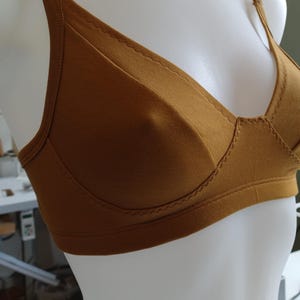 May include: A mustard-colored bra with adjustable straps and a classic design. The bra is displayed on a mannequin, showcasing its shape and construction. The fabric appears to be a soft, solid material with subtle stitching details.