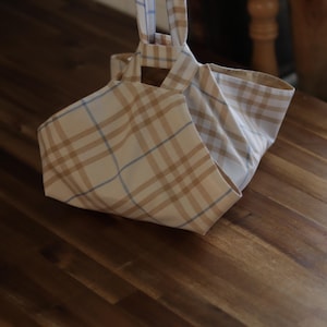 May include: A beige and brown plaid tote bag with matching handles. The bag is made of fabric and is folded open, revealing the interior. The bag is hanging from a wooden rod.