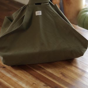 May include: Olive green fabric tote bag with a rectangular base and integrated handles. A small, rectangular label is sewn onto the front. The bag is sitting on a wooden surface.