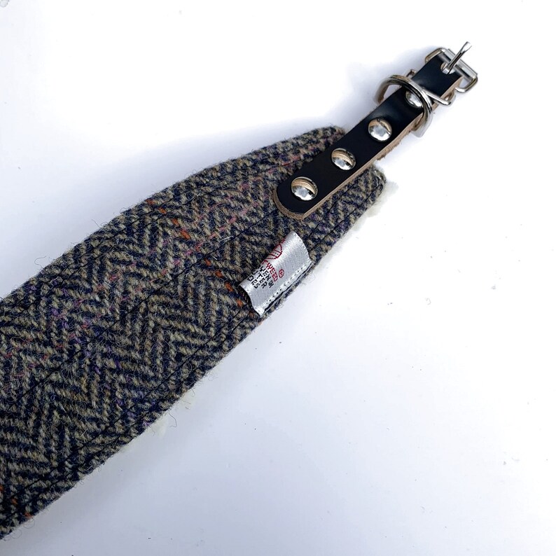 May include: A tweed dog collar with a black leather strap and silver metal hardware. The collar has a white label with the text "TWEED" and "HAND MADE IN SCOTLAND".
