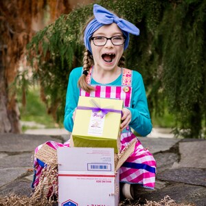 May include: A young girl wearing a blue headband, glasses, and a pink and white gingham dress is opening a yellow box with a purple ribbon. The box says "Fairy" on it. The girl is sitting on a stone surface with brown packing paper.