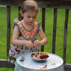 May include: A young child wearing a patterned dress is focused on a small bowl filled with miniature decorative items. The bowl sits on a galvanized metal surface, with a wooden railing and green grass in the background.