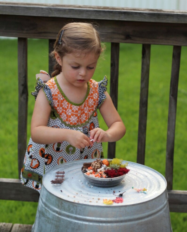 May include: A young child wearing a patterned dress is focused on a small bowl filled with miniature decorative items. The bowl sits on a galvanized metal surface, with a wooden railing and green grass in the background.