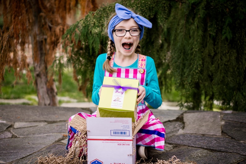 May include: A young girl wearing a blue headband, glasses, and a pink and white gingham dress is opening a yellow box with a purple ribbon. The box says "Fairy" on it. The girl is sitting on a stone surface with brown packing paper.