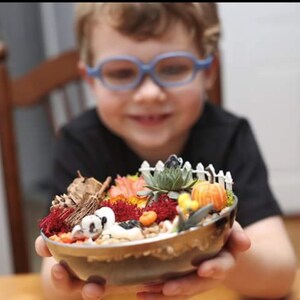 May include: A decorative miniature autumn-themed terrarium in a small metal bowl. The terrarium features a white picket fence, a small pumpkin, and various plants and decorative elements. The terrarium is held by a person.