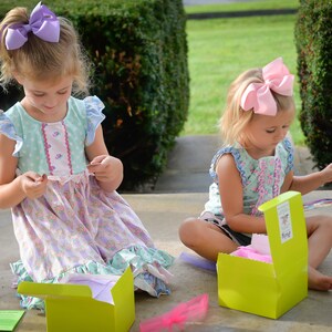 May include: Two young girls are sitting on a porch, opening colorful boxes. One girl is wearing a light blue and pink dress with a purple bow in her hair. The other girl is wearing a light blue and white dress with a pink bow in her hair. Both girls are holding small cards with text.
