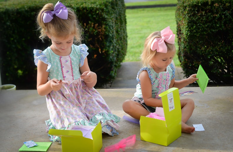May include: Two young girls are sitting on a porch, opening colorful boxes. One girl is wearing a light blue and pink dress with a purple bow in her hair. The other girl is wearing a light blue and white dress with a pink bow in her hair. Both girls are holding small cards with text.