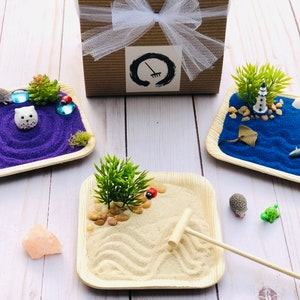 May include: Miniature zen gardens with various themes. One features purple sand with an owl figurine, another has blue sand with a lighthouse, and the third has tan sand with a rake. A gift box with a white bow is in the background.