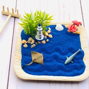 May include: A miniature beach scene with a blue lighthouse, a pink flamingo, a stingray, a dolphin, and a seashell on a blue sand base. The scene is surrounded by a brown tray with a wooden rake.