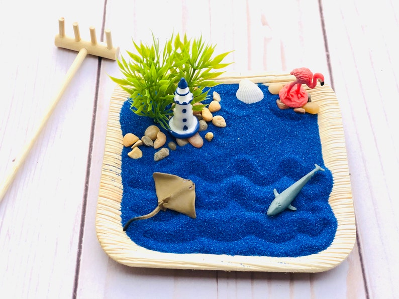 May include: A miniature beach scene with a blue lighthouse, a pink flamingo, a stingray, a dolphin, and a seashell on a blue sand base. The scene is surrounded by a brown tray with a wooden rake.