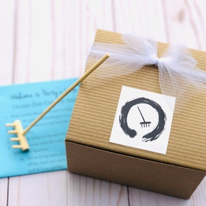 May include: A brown cardboard box with a white ribbon and a white tulle bow. A sticker with a black and white illustration of a rake inside a circle is on the front of the box. The box is sitting on a white wooden surface.