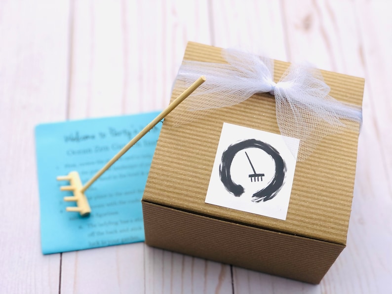 May include: A brown cardboard box with a white ribbon and a white tulle bow. A sticker with a black and white illustration of a rake inside a circle is on the front of the box. The box is sitting on a white wooden surface.