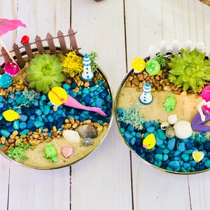May include: Two miniature beach scenes in round containers. Each scene features a mermaid, a small lighthouse, a succulent plant, and a variety of colorful stones and shells. One scene has a pink umbrella and the other has an orange umbrella.