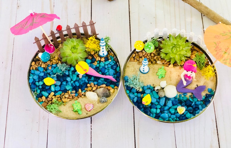 May include: Two miniature beach scenes in round containers. Each scene features a mermaid, a small lighthouse, a succulent plant, and a variety of colorful stones and shells. One scene has a pink umbrella and the other has an orange umbrella.